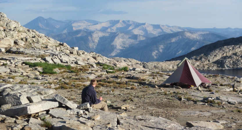 A person sits on rocky ground near a small tent set up at high elevation overlooking a mountainous area. 